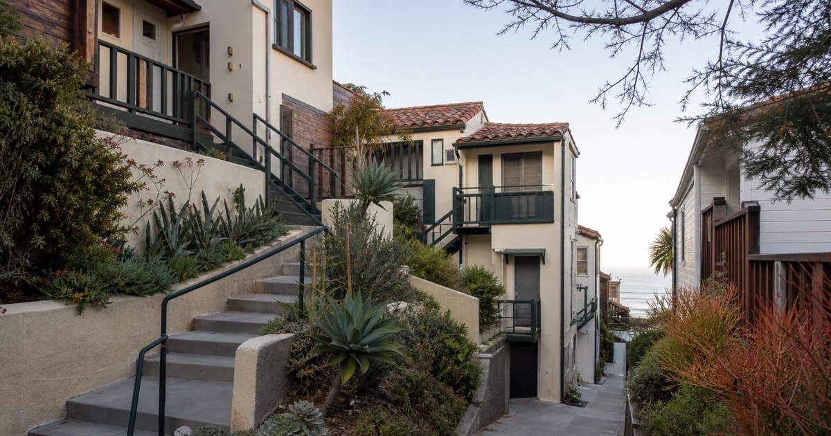 Steep Laguna Beach rental lane with cream stucco homes, green railings, and a distant ocean strip.